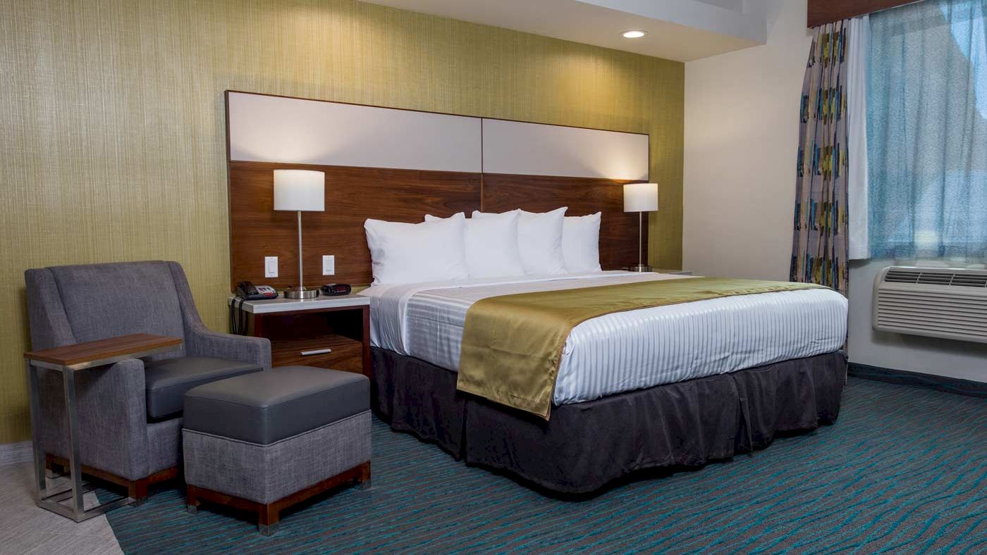A modern hotel room with a large bed, lamps, an armchair with a footrest, a side table, and an air conditioner by the window.