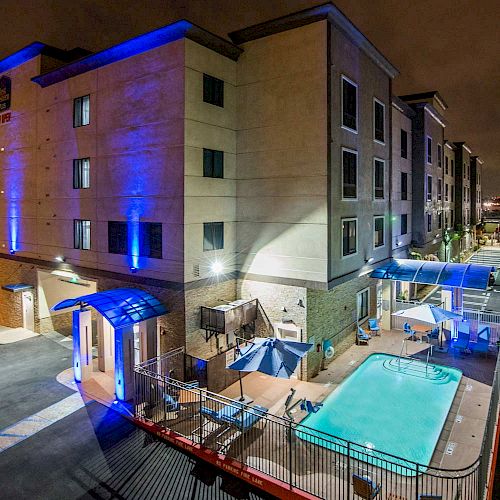 The image shows a multi-story hotel building at night, lit with blue lights, featuring an outdoor swimming pool and several lounge chairs.