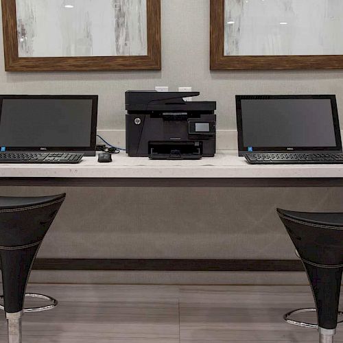 The image shows a workspace with two laptops, a printer in between, and two bar stools in front of a desk, with picture frames on the wall.