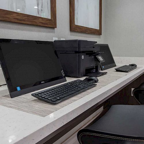 The image shows a business center with two computers, a printer, and two chairs on a countertop. The room has a clean, modern look.