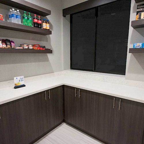 The image shows a pantry with shelves holding drinks, snacks, and other items, along with cabinets below the countertop and a small refrigerator.