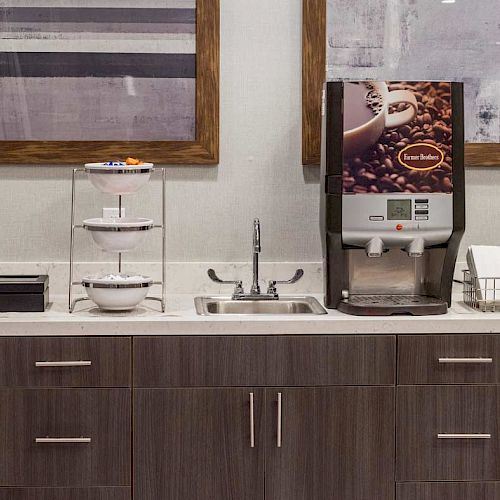 The image shows a coffee station with a coffee machine, cups, utensils, and storage cabinets below, along with framed art on the wall behind.