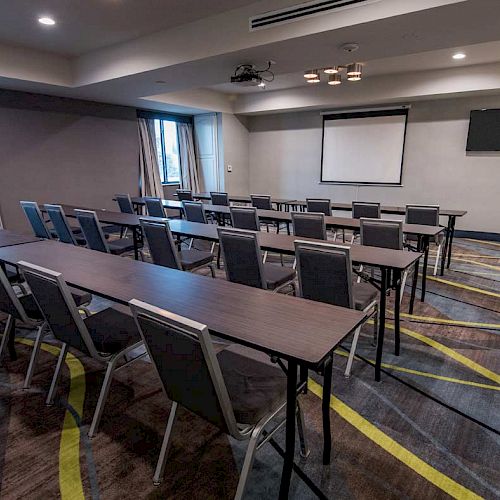 This image shows a conference room with rows of chairs and tables facing a screen and a TV, well-lit with ceiling lights and windows.