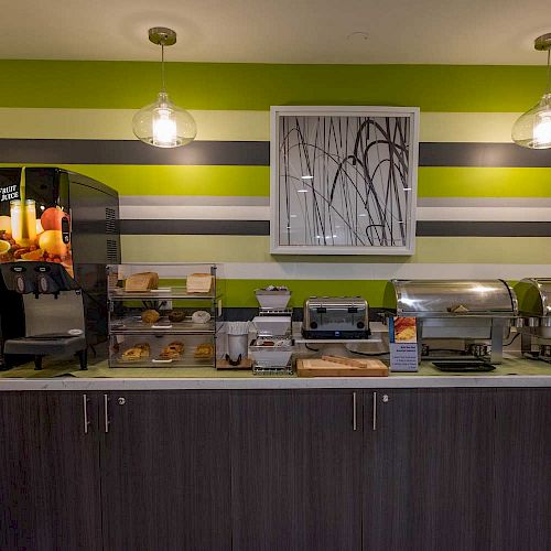 A modern breakfast buffet setup with a coffee machine, cereal dispensers, pastries, and chafing dishes on a counter below striped walls.