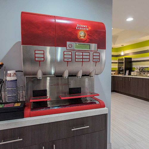 The image shows a breakfast area with a red beverage dispenser, cups, and condiments on the counter. Another counter with additional machines is in the background.