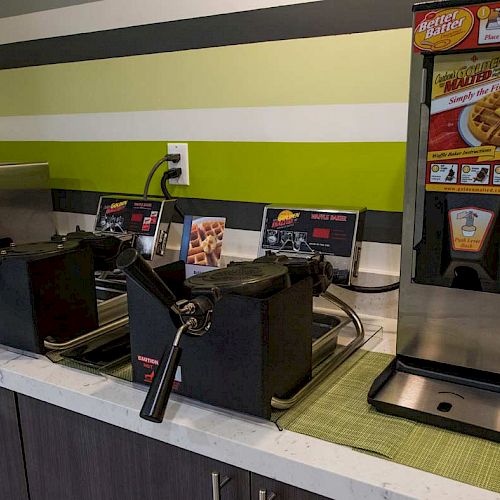 A breakfast station with waffle makers, a waffle batter dispenser, a microwave, and syrup containers on a counter in a green-striped room.