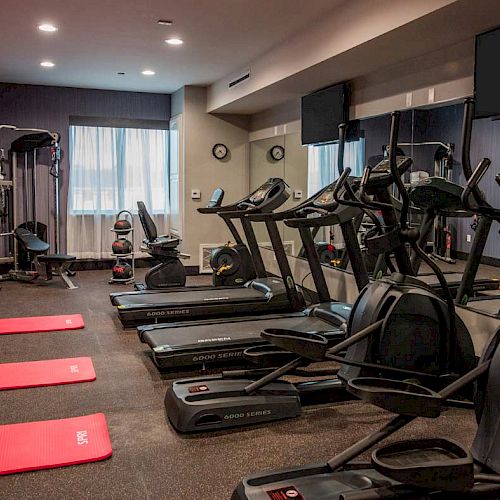 This image shows a well-equipped indoor gym with treadmills, elliptical machines, red exercise mats, weight machines, and mirrors on the walls.