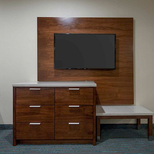 The image shows a modern room setup with a mounted flat-screen TV, wooden dresser with drawers, and a small side table, all against a beige wall.