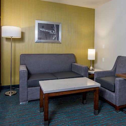 The image shows a hotel room with a grey sofa, armchair, wooden coffee table, two lamps, a framed picture, and a bed partially visible on the left.