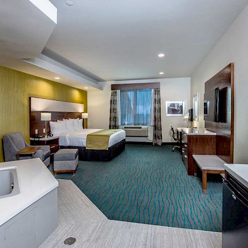 The image shows a hotel room with a bed, a seating area, a TV, a microwave, a mini-fridge, and a hot tub in the corner, featuring modern decor.