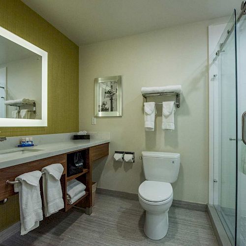 The image shows a modern bathroom with a sink, mirror, toilet, glass-enclosed shower, and neatly arranged towels on shelves and racks.