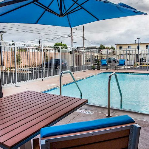 An outdoor pool area with blue umbrellas, chairs, and a wooden table, surrounded by a white metal fence. A few buildings are in the background.