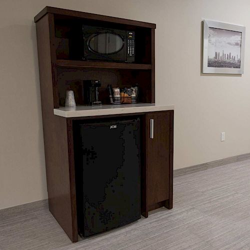 A kitchenette setup with a microwave, mini-fridge, coffee maker, and some condiments, against a wall with a framed picture.