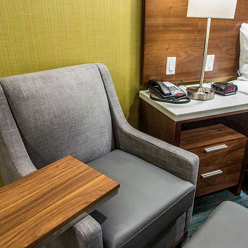 The image shows a gray armchair with a wooden side table, next to a nightstand with a lamp, phone, and alarm clock.