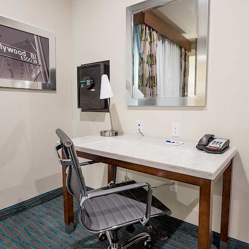 A small office area with a desk, chair, lamp, phone, and notepad. There's a framed picture on the wall and a mirror reflecting a window.