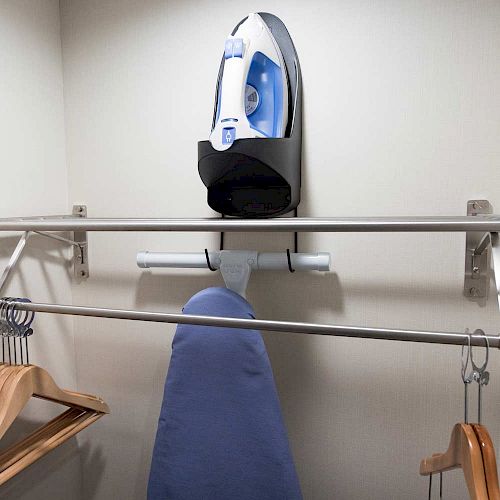 The image shows a closet with an iron on a wall-mounted holder, an ironing board, a metal rod with hangers, and an additional metal rod above it.