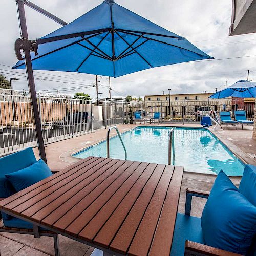 The image shows an outdoor pool area with a wooden table, blue chairs, and umbrellas, surrounded by a fence and buildings in the background.