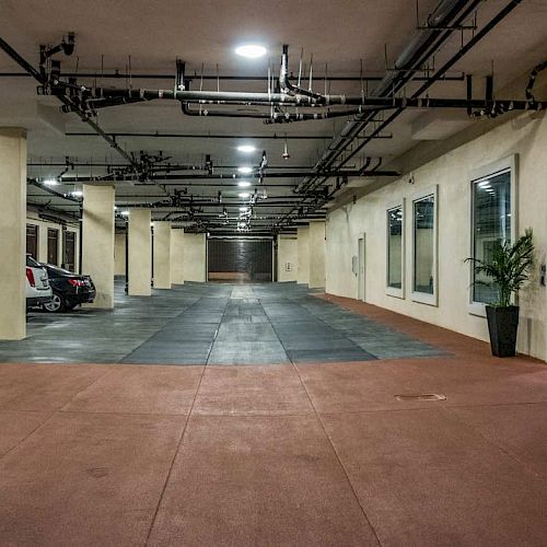 An indoor parking area with a few parked cars, columns, overhead piping, potted plants, and an entrance to a building.