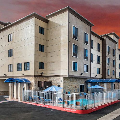 A multi-story hotel building with blue awnings and a fenced outdoor pool area is shown, set against a vibrant sunset sky.