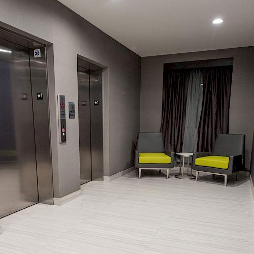 The image shows an elevator lobby with two elevators, two chairs with green cushions, a small table, a curtain, and a wall-mounted phone.