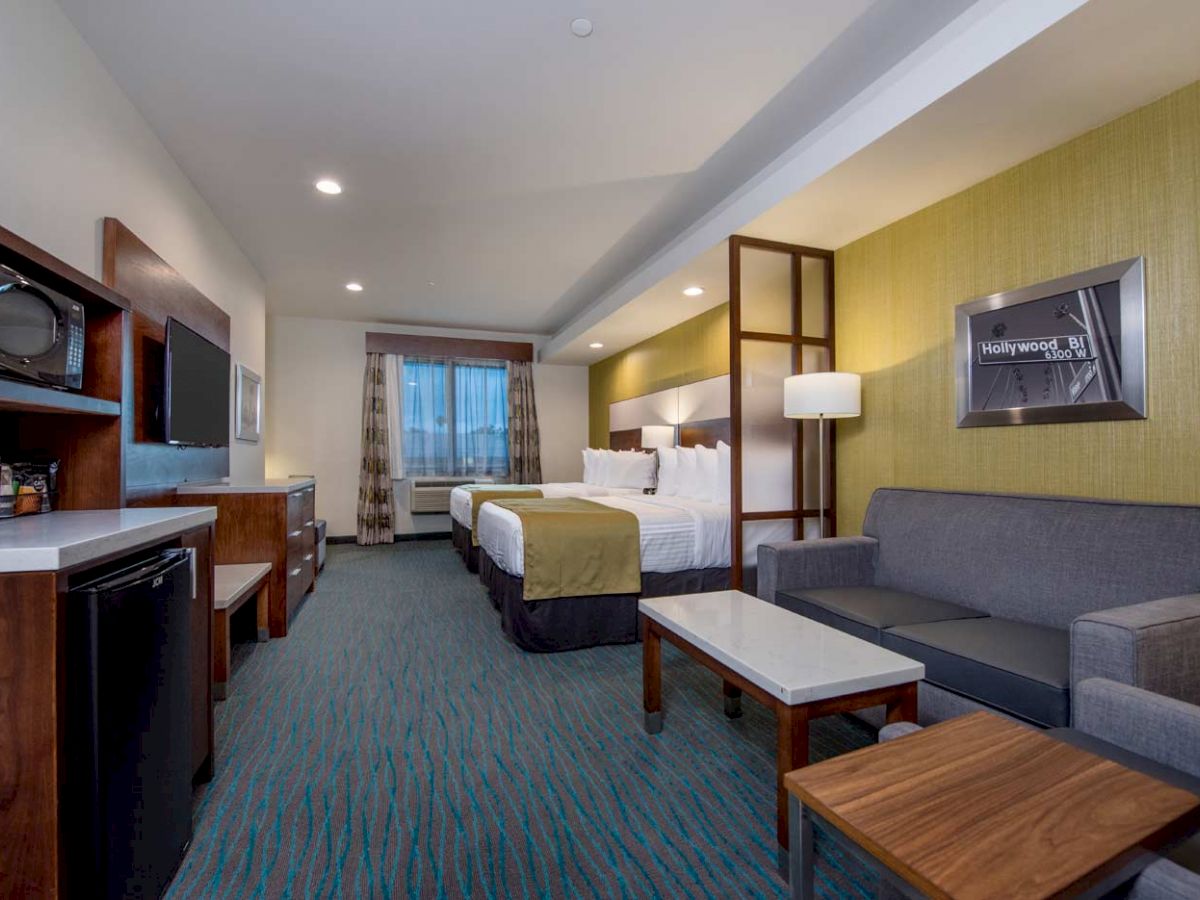 The image shows a hotel room with two beds, a gray sofa, wooden tables, a TV, a microwave, and a mini-fridge.