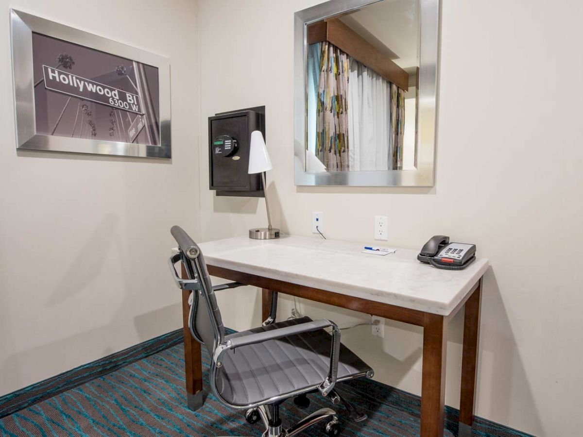 The image shows a small office or hotel room workspace with a desk, chair, phone, and desk lamp. A framed picture and mirror adorn the walls.