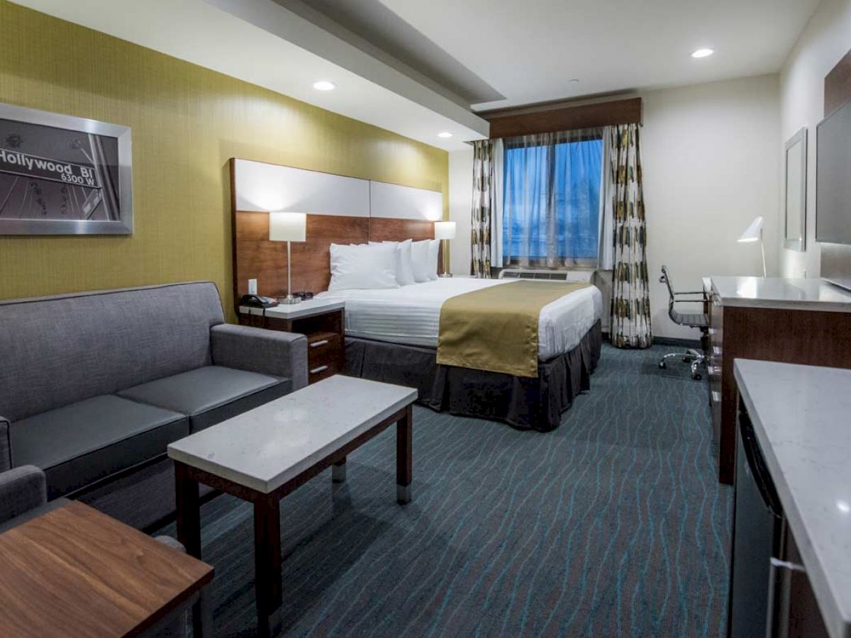 A modern hotel room with a bed, sofa, coffee table, desk, and chair. There's a window with curtains and a framed picture on the wall.