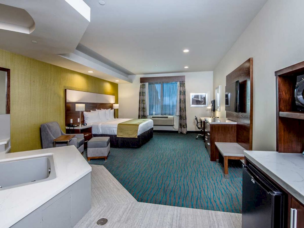 The image shows a hotel room with a bed, sitting area, desk, TV, microwave, mini-fridge, and a jacuzzi tub.