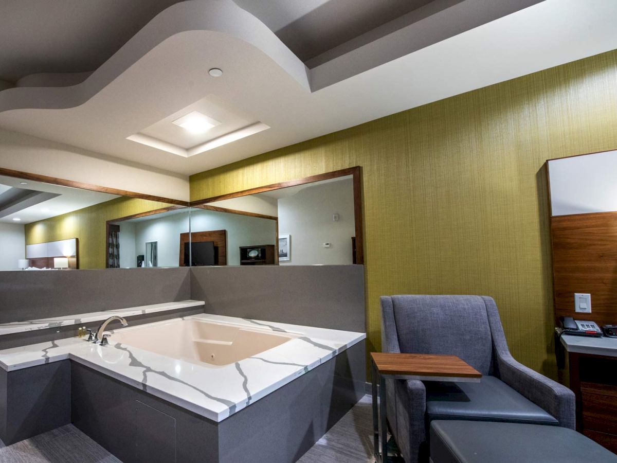 The image shows a modern hotel room with a large bathtub, a grey armchair with a small table, and warm lighting.