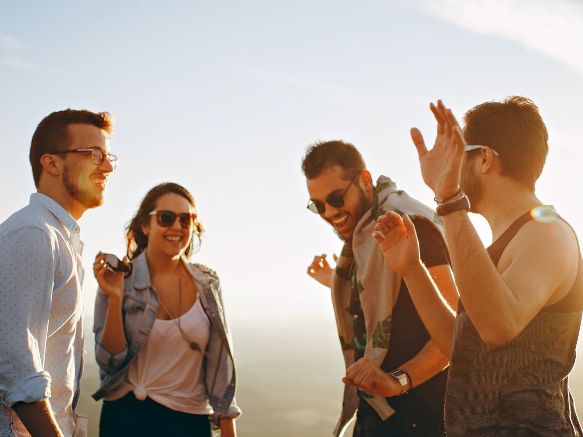 Four people standing outdoors, smiling and chatting with each other in a cheerful, sunlit setting.