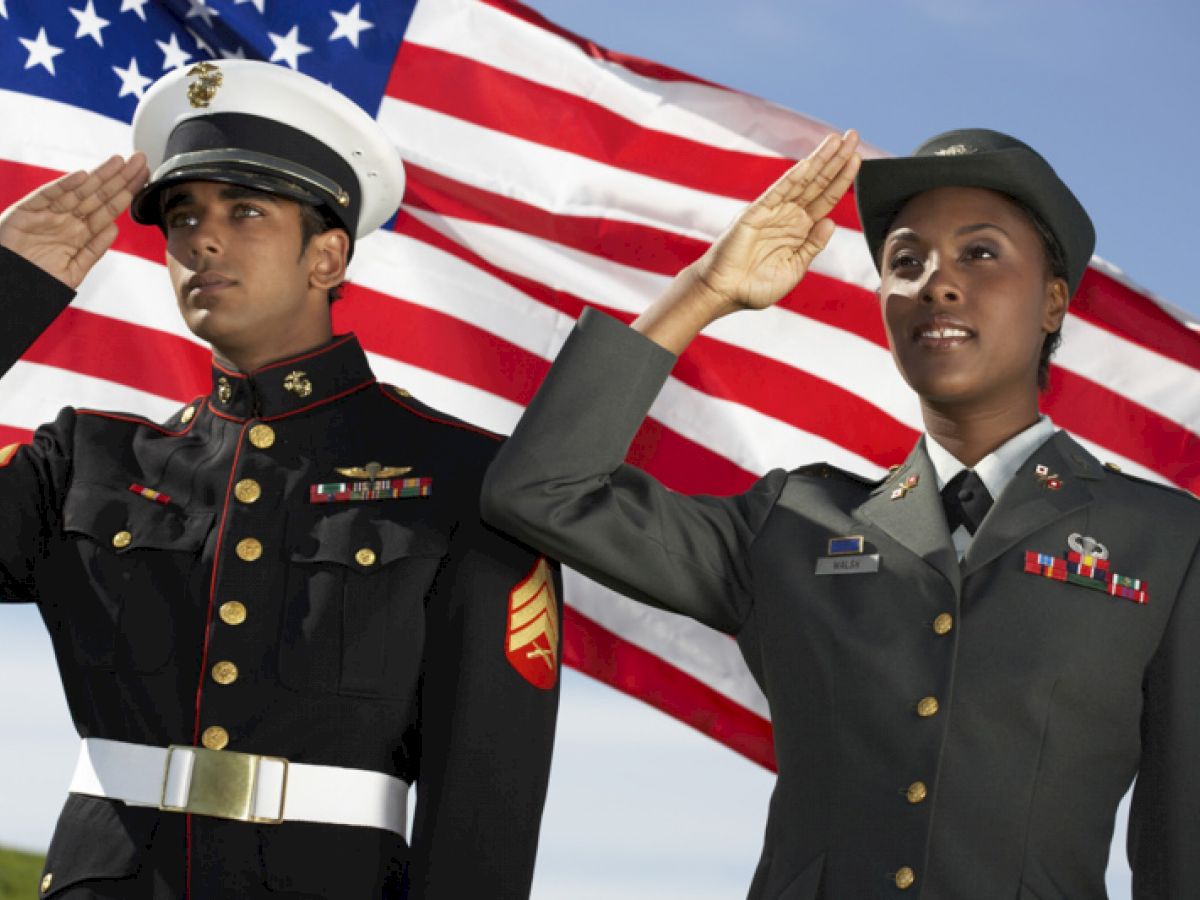 Two uniformed military personnel are saluting with an American flag in the background, standing outdoors under a clear sky.