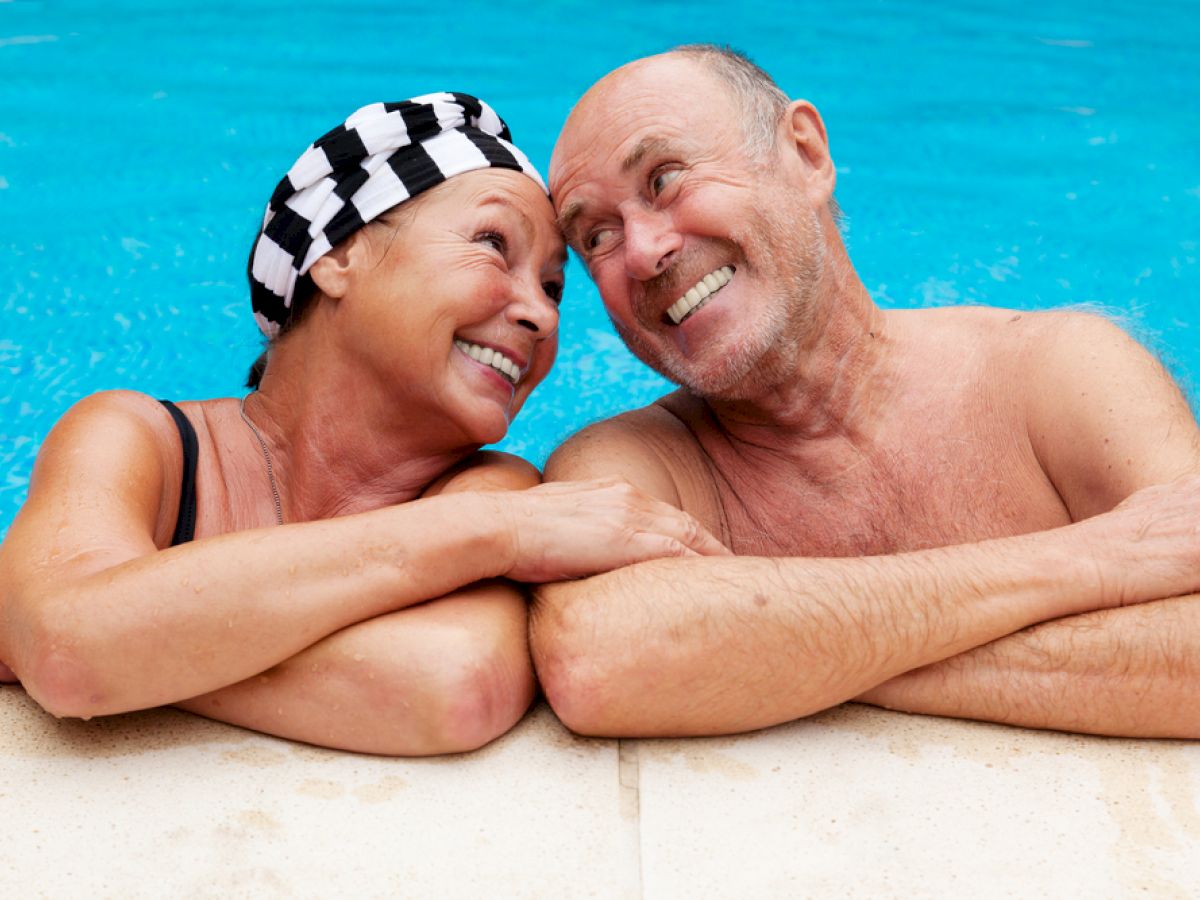 An elderly couple is smiling at each other while relaxing on the edge of a swimming pool, enjoying a sunny day outdoors.