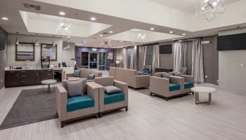 A modern lounge area featuring several beige and teal armchairs, coffee tables, a counter with refreshments, and contemporary lighting fixtures.