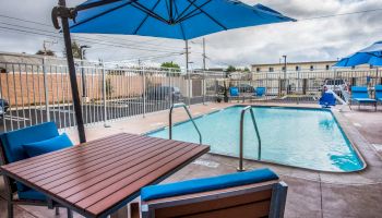 An outdoor swimming pool area features blue umbrellas, lounge chairs, and a wooden table with chairs, surrounded by a white fence.