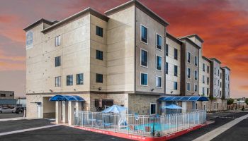 The image shows a multi-story beige building with blue awnings and a small fenced pool area outside. The sky is a vibrant orange and red at sunset.