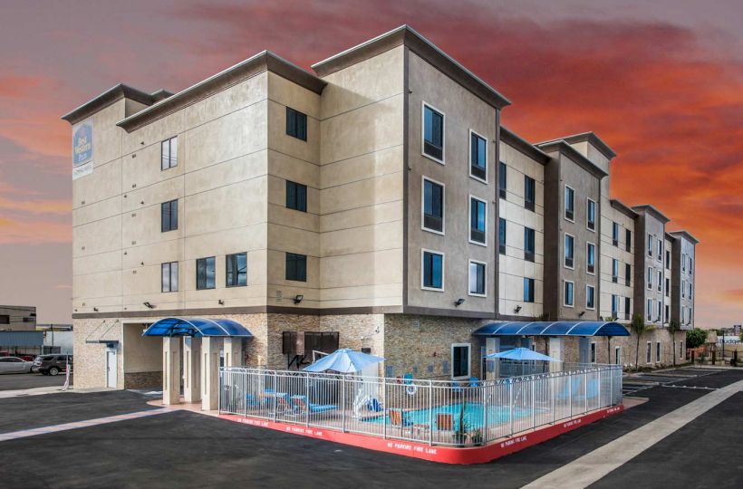 The image shows a multi-story beige building with blue awnings and a small fenced pool area outside. The sky is a vibrant orange and red at sunset.