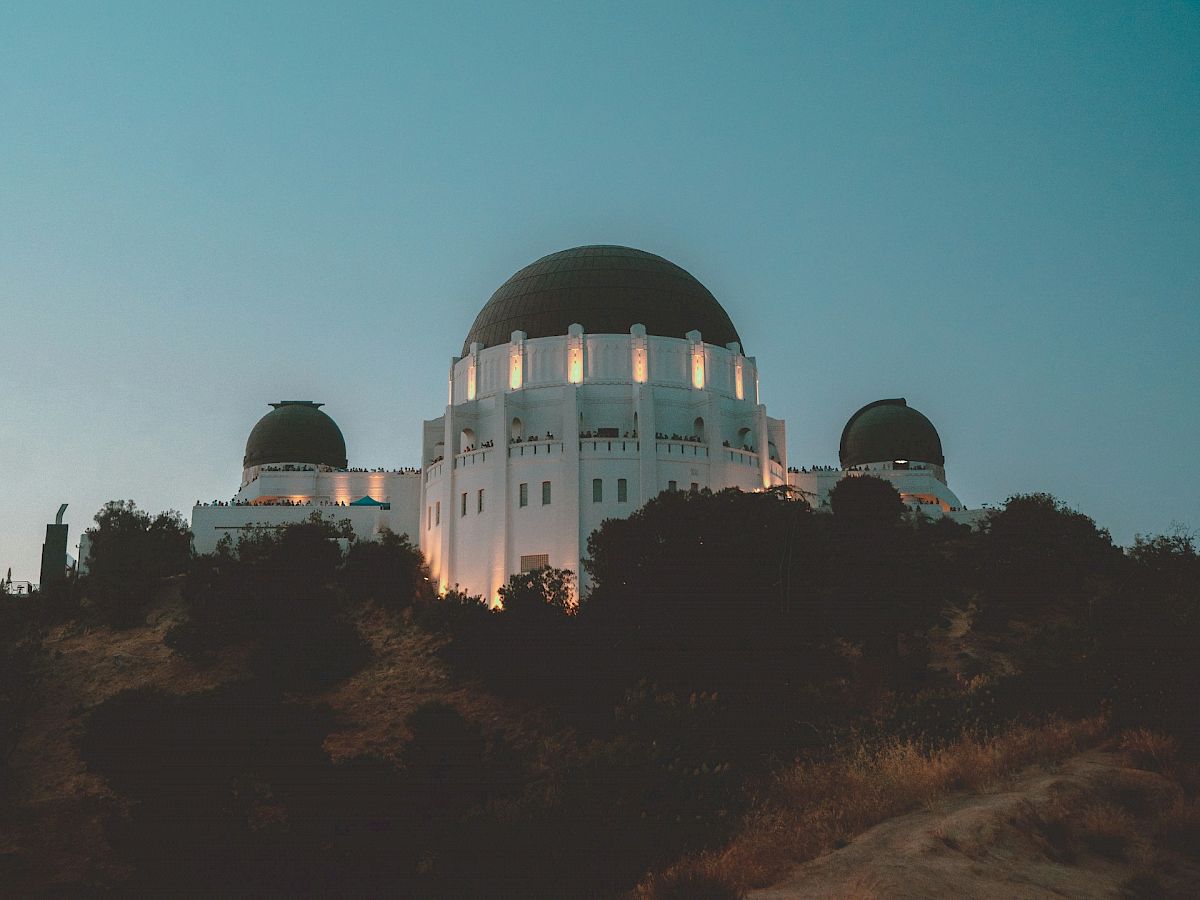 The image shows a large white observatory with a central dome and two smaller domes, set against a twilight sky, surrounded by trees.