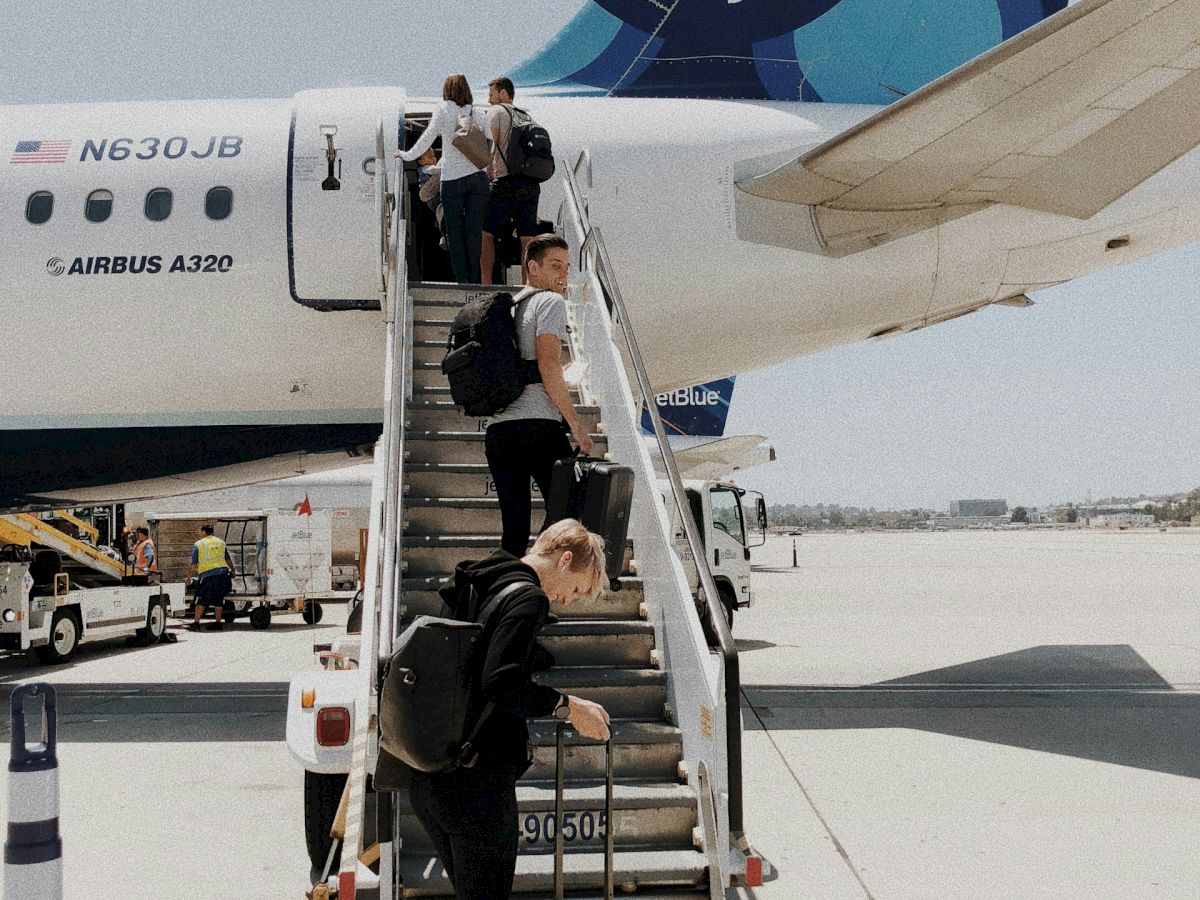 People are boarding a jetBlue airplane via stairs on the airport tarmac, carrying luggage and preparing to enter the aircraft.