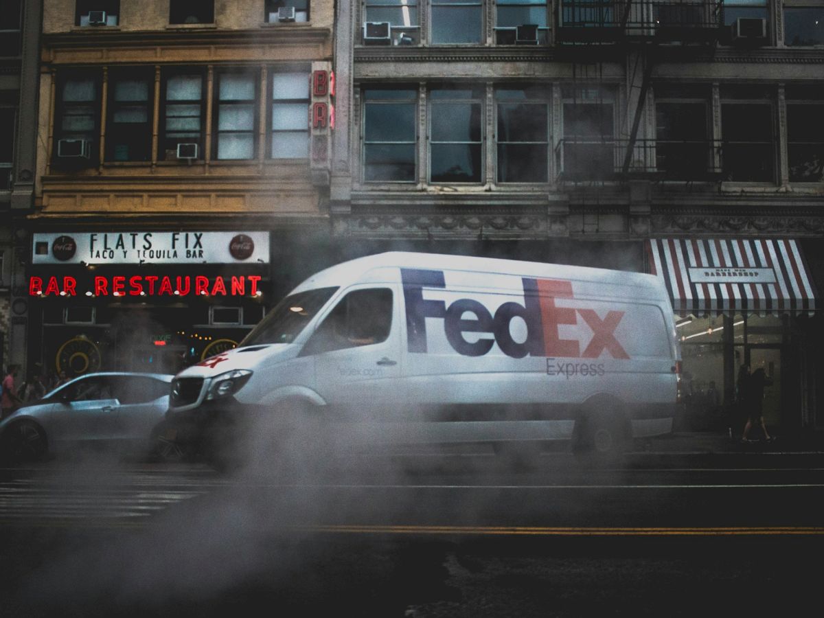 A white FedEx van is parked on a street in front of buildings with various signs, with steam rising from the ground around it.