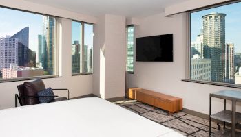 A modern hotel room with large windows showcasing a city skyline, a wall-mounted TV, armchair, and minimalistic furniture.