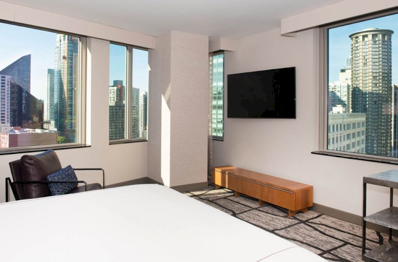 A modern hotel room with large windows showcasing a city skyline, a wall-mounted TV, armchair, and minimalistic furniture.