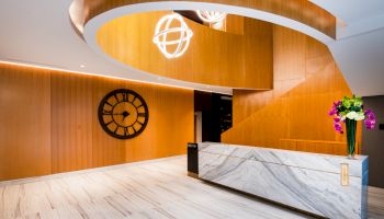 Modern lobby with a marble reception desk, wooden spiral staircase, spherical light fixture, large wall clock, and a vase with colorful flowers.