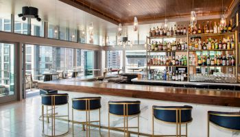 The image shows a modern bar with high stools, a well-stocked liquor shelf, and large windows with a view of city buildings.