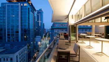 The image shows a modern cityscape viewed from a stylish balcony at dusk. The balcony features seating and tables, and large windows.