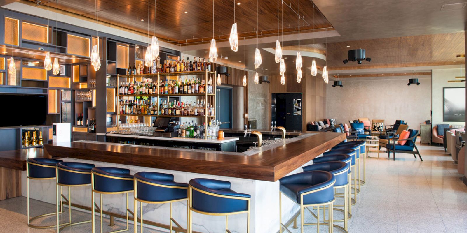 The image depicts a modern bar with wooden accents, blue bar stools, hanging lights, and a well-stocked bar counter. The setting is elegant and inviting.