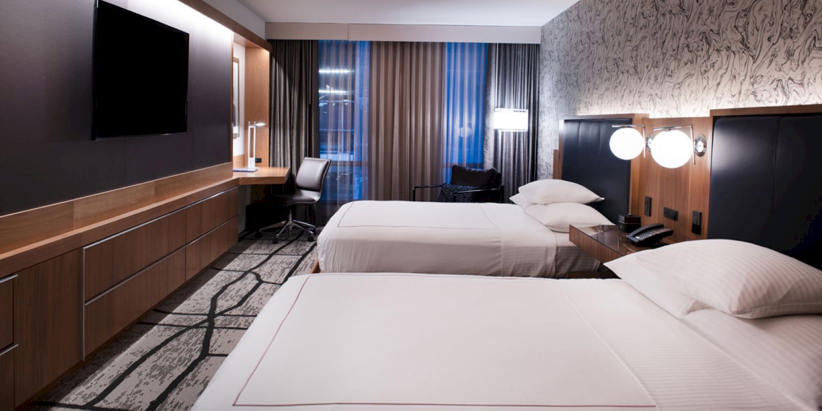 A modern hotel room with two neatly made beds, a wall-mounted TV, a desk with a chair, and stylish lighting fixtures.