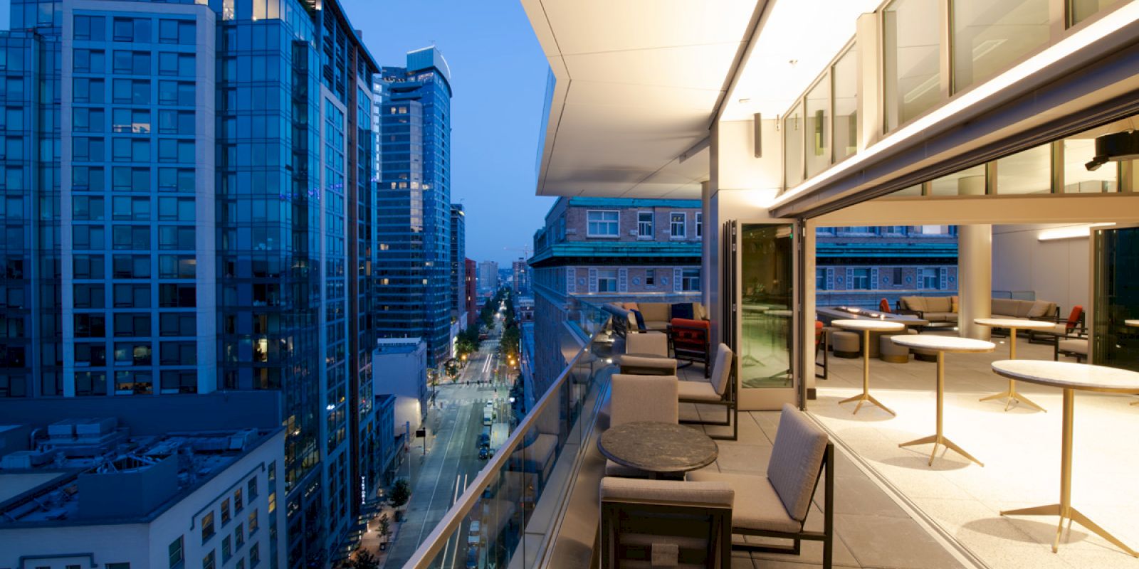 A modern rooftop terrace at dusk with tables and chairs, adjacent to a city street lined with tall buildings.