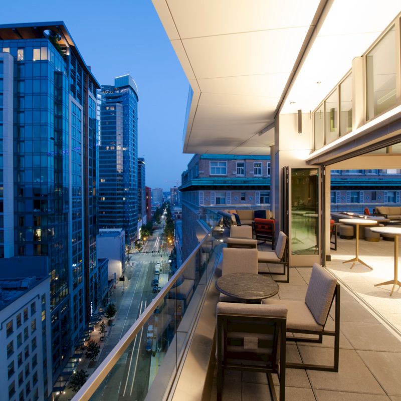 The image shows a modern cityscape with tall buildings and a balcony overlooking the street, featuring outdoor seating and tables.