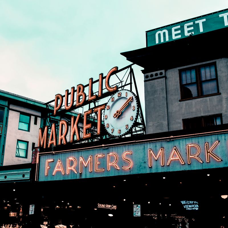 The image shows an iconic sign for a Public Market and Farmers Market, likely indicating a well-known marketplace, with buildings in the background.