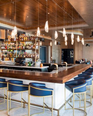 A modern, stylish bar with a wooden counter, blue chairs, a well-stocked liquor shelf, hanging lights, and a seating area in the background.